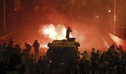 A riot police officer, on a armoured personnel carrier surrounded by anti-Mursi protesters (foreground), fires rubber bullets