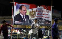 People walk in front of a banner supporting President Abdel Fattah al-Sisi's re-election hanging in a clothes market near the canal of Port Said, Egypt March 18, 2018. PHOTO BY REUTERS/Mohamed Abd El Ghany