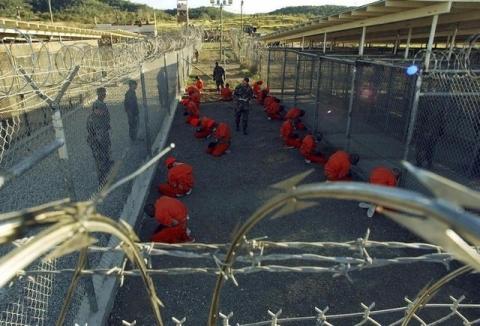 Detainees in orange jumpsuits sit in a holding area under the watchful eyes of military police during in-processing to the temporary detention facility at Camp X-Ray of Naval Base Guantanamo Bay, January 11, 2002. PHOTO BY REUTERS/Stringer