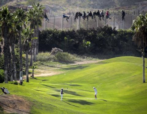 A golfer hits a tee shot as African migrants sit atop a border fence during an attempt to cross into Spanish territories between Morocco and Spain's north African enclave of Melilla, October 22, 2014. REUTERS/Jose Palazon